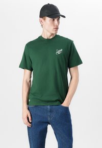 T-shirt en coton vert à manches courtes, arborant un logo Lacoste blanc sur la poitrine. Porté avec un jean en denim bleu et une casquette noire.