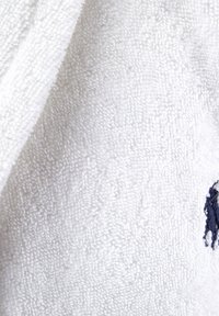 Accappatoio bianco in morbido tessuto di spugna con texture. Presenta un piccolo logo blu navy sul davanti. Tessuto morbido, peloso e assorbente.