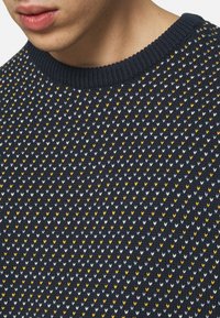 Stickad tröja med en marinblå bas och ett mönster av små, repetitiva gula, vita och ljusgrå pilar. Ribbat rundhalsat nacke.