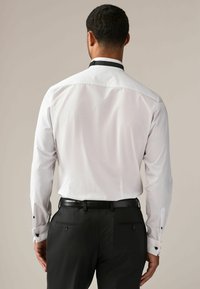 Witte blouse met lange mouwen en een zwarte kraag, met knopen op de mouwen en een soepele, op maat gemaakte pasvorm zichtbaar vanaf de achterkant. Zwarte riem en broek.