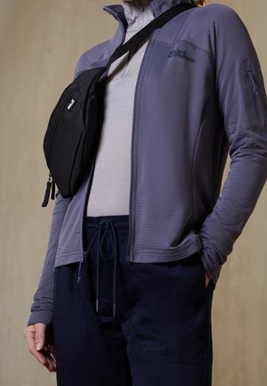 Person trägt eine graue Jacke mit Reißverschluss, hellgraues Hemd, marineblaue Hose mit Kordelzug und eine schwarze Umhängetasche quer über der Brust.