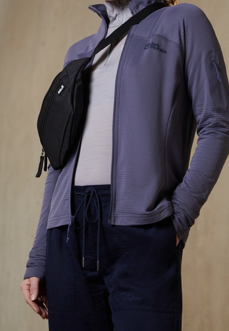 Persona vestida con chaqueta gris con cremallera, camisa gris clara, pantalones azul marino con cordón y un bolso negro cruzado sobre el pecho.
