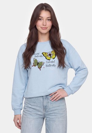 Jonge vrouw met lang bruin haar draagt een lichtblauwe sweatshirt met kleurrijke vlinderafdrukken en lichtblauwe spijkerbroek, hand op de heup.