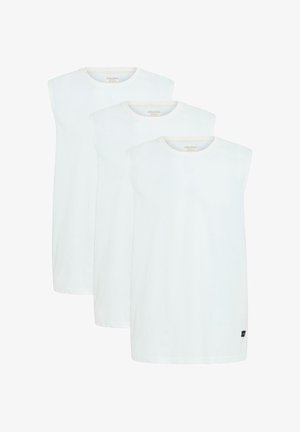 Jonge man met kort donker haar, gekleed in een mouwloos wit shirt, losse zwarte jeans en witte sneakers met beige strepen, staand met het gezicht naar voren.