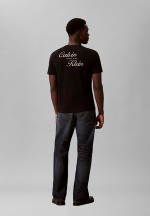 Mężczyzna zwrócony tyłem, ubrany w czarną koszulkę Calvin Klein z logo na plecach oraz ciemnoniebieskie dżinsy na jednolitym szarym tle.