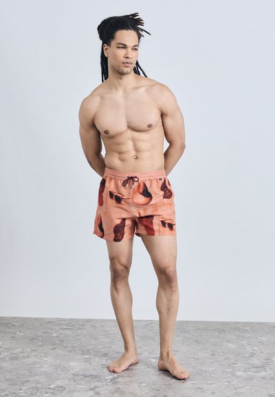 Homme torse nu avec des dreadlocks portant un short de bain orange avec des motifs abstraits noirs et rouge foncé, debout pieds nus sur un sol gris.