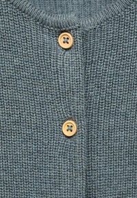 Pleteni cardigan v sivo-modri barvi z rebrasto teksturo, ki ima dva lesena gumbka. Ovratnik je krogu in se razteza spredaj.