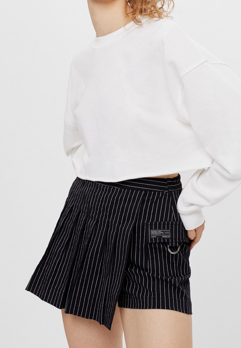 Sweat-shirt blanc court associé à un short noir à fines rayures avec plis, une poche avec une étiquette et un anneau métallique en accent.