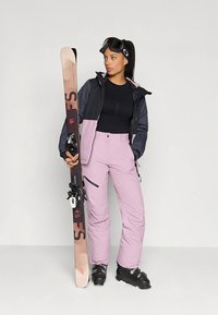 Skidkläder med en svart och rosa jacka, ljusrosa byxor, svart topp och svarta skidpjäxor, håller skidor med en lila och beige design.