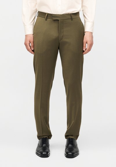 Άνδρας που φοράει ελαιί πράσινο παντελόνι φόρμας, λευκό πουκάμισο και μαύρα αθλητικά παπούτσια, στέκεται μπροστά σε απλό φόντο.