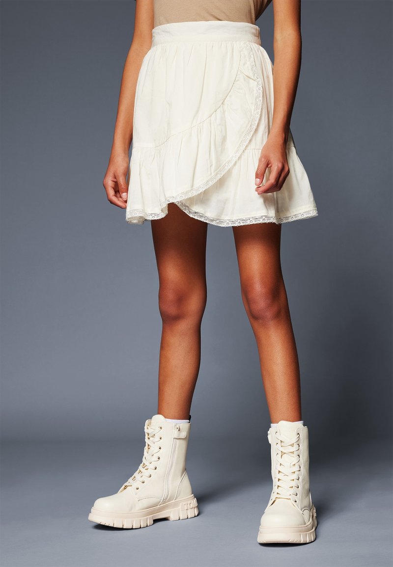 Femme portant une jupe portefeuille blanche avec bordure en dentelle et des bottes de combat beige à lacets, debout devant un fond gris uni.