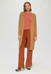Lange tan cardigan met een capuchon over een roestkleurig gestreept shirt, gecombineerd met roestkleurige wijde corduroy broek en meerkleurige sneakers.