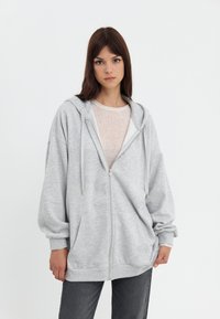 Sweat à zip couleur gris clair avec une coupe spacieuse, une texture douce, une capuche ajustable et de grandes poches avant. Porté par-dessus un haut clair et un jean gris.