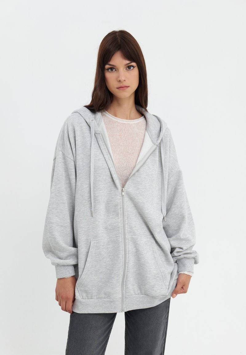 Sweat à zip couleur gris clair avec une coupe spacieuse, une texture douce, une capuche ajustable et de grandes poches avant. Porté par-dessus un haut clair et un jean gris.