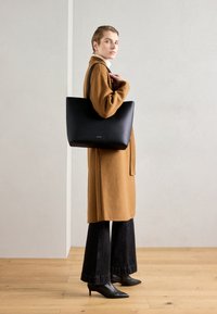 Black leather tote bag with a soft finish, featuring a structured shape and contrast stitching, carried against a light gray background.