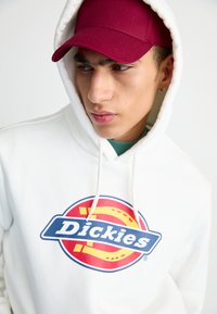 White hooded sweatshirt with a large blue and red circular "Dickies" logo. Paired with a maroon cap, featuring a smooth fabric texture.