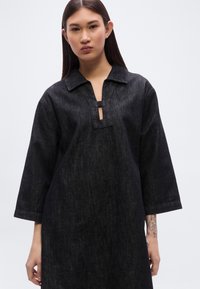 Μαύρο τουνίκ φόρεμα από τζιν με μανίκια τριών τετάρτων, γιακά και μικρή οβάλ εγκοπή στο λαιμό. Λείο ύφασμα και άνετη εφαρμογή.