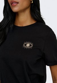 Femme portant un t-shirt noir avec un motif d'œil brodé beige sur le côté gauche de la poitrine et des boucles d'oreilles en créoles dorées.