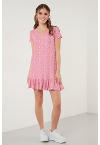 Korte roze jurk met een v-hals en korte mouwen, met een gerimpelde zoom en een wit bloemenpatroon over de hele jurk. Draagt witte sneakers.
