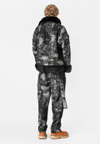 Persona con chaqueta y pantalones texturizados en negro y gris con puños de piel, botas naranjas y detalles de etiquetas en los pantalones, de pie de espaldas.