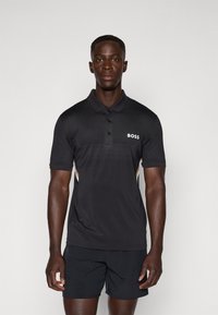 Polo shirt noir avec un design texturé, trois boutons, logo sur la poitrine et bandes latérales contrastées. Porté avec un short noir.