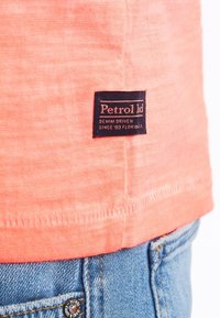 T-shirt corallo con etichetta sul fondo che riporta "Petrol ltd" e dettagli di cucitura, abbinato a jeans in denim blu con un accentuato bottone circolare.