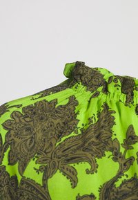 Grön blus med invecklade bruna blommönster, med en samlad halsringning och strukturerad tyg. Fokus på design detaljer och färgkontrast.