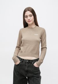 Bež skrajšan pulover z rebrastimi manšetami in robom, z belim napisom "2ND DAY" na prsih. Kombiniran z visokim pasom črnih jeans hlač.