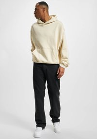 Beige oversized hoodie with a kangaroo pocket, paired with black straight-leg jeans and white sneakers. Soft texture, casual design.
