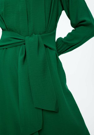 Grünes Kleid aus einem weichen, strukturierten Stoff. Es hat lange Ärmel, einen gebundenen Gürtel in der Taille und einen fließenden Rock.