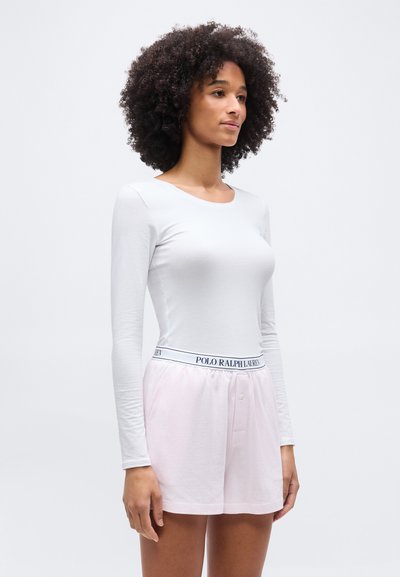 Γυναίκα με σγουρά μαλλιά που φοράει λευκό μακρυμάνικο τοπ και ροζ σορτς με ζώνη Polo Ralph Lauren, στέκεται μπροστά σε απλό φόντο.