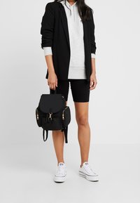 Czarny nylonowy plecak ze złotymi elementami; posiada klapę, przednie kieszenie oraz regulowane paski. Nosi się go z szarą bluzą z kapturem i czarnymi szortami.