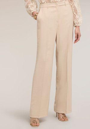 Femme portant un pantalon large beige taille haute, des sandales à talons dorées à brides, un top en dentelle à manches longues et des bracelets dorés au poignet gauche.