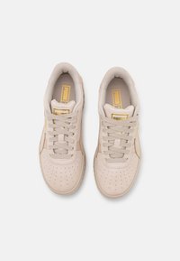 Baskets de sport beiges avec un design perforé, tige en suède texturé, lacets plats, et accents dorés sur la languette et les côtés.