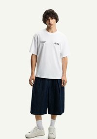 White cotton t-shirt with short sleeves and black text, paired with dark denim shorts. White sneakers and white socks.