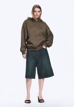 Femme portant un sweat à capuche olive oversize, un short en jean large et foncé arrivant aux genoux, et des sandales à talons ouvertes noires, debout devant un fond blanc.
