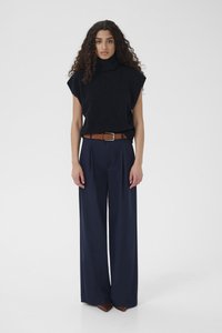 Kobieta o kręconych włosach, ubrana w czarny bezrękawnik z golfem, granatowe szerokie spodnie, brązowy pasek i brązowe szpiczaste buty, stoi na białym tle.