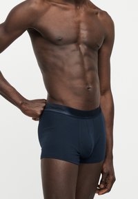 Μυώδης κορμός ενός σκουρόχρωμου άνδρα που φοράει σκούρο μπλε μποξεράκια Calvin Klein, με το ένα χέρι να ρυθμίζει τη μέση.