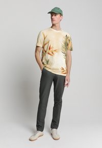 Korte mouwen beige T-shirt met tropische oranje en groene plantprints, gecombineerd met donkergrijze broek en lichte sneakers, afgemaakt met een groene pet.