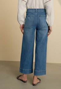 Jeans de mezclilla azul de pierna ancha con una cintura alta, con dobladillos acabados y dos bolsillos traseros, combinados con una blusa blanca ligera y unas sandalias estampadas.