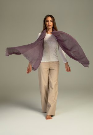 Leichter lila Schal mit ausgefransten Kanten, leger über einem weißen Langarmshirt getragen, kombiniert mit cremefarbenen, locker sitzenden Hosen.