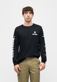 Νεαρός άνδρας που φοράει μαύρο μακρυμάνικο πουκάμισο Converse με λευκά λογότυπα στο στήθος και στα μανίκια, συνδυασμένο με χακί παντελόνι και μαύρη ζώνη.