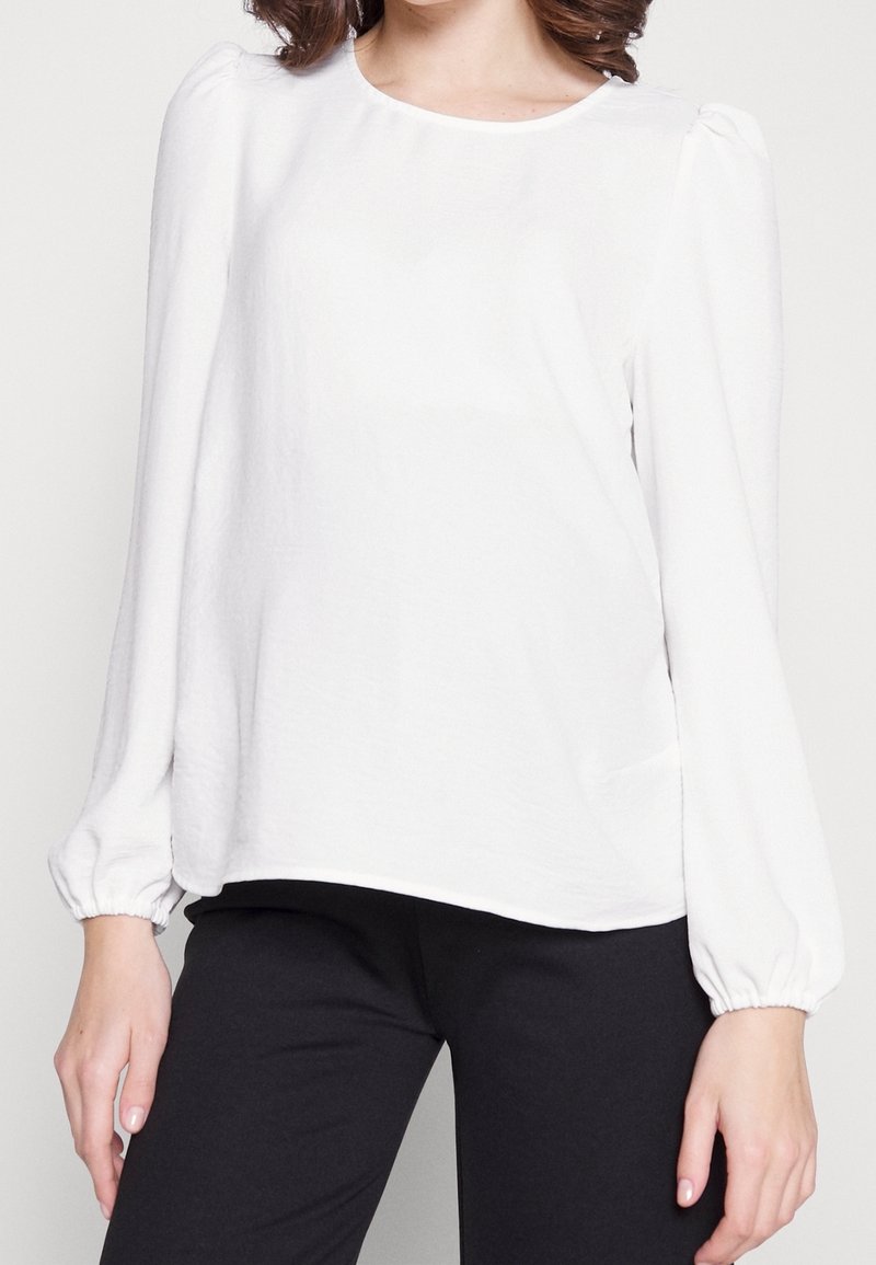Femme portant un chemisier blanc à manches longues avec des épaules bouffantes et des poignets élastiques, associé à un pantalon noir sur un fond uni.