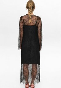 Černé krajkové šaty s dlouhými průhlednými rukávy a asymetrickým spodním lemem. Materiál má složité květinové vzory a hladkou spodní vrstvu.