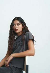 Un t-shirt en coton gris avec des manches courtes et une coupe décontractée, orné d'un motif en texte noir. Porté avec un jean foncé, assis contre un fond clair.