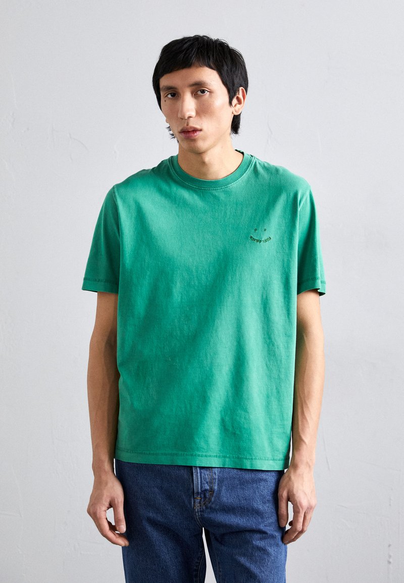 Πράσινο βαμβακερό T-shirt με στρογγυλή κοπή και κοντά μανίκια, διαθέτοντας μια μικρή κεντητή σχεδίαση στην αριστερή πλευρά του στήθους. Φοριέται με μπλε τζιν παντελόνι.