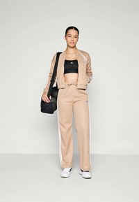 Fato de treino atlético bege com riscas brancas, apresentando uma jaqueta com fecho e calças com cordão. Usado com um top cropped preto e uma mala preta.