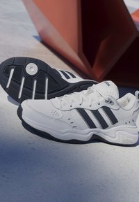 Baskets blanches Adidas avec des bandes noires, montrant le côté et la semelle, placées sur une surface claire à côté d'une sculpture géométrique marron.