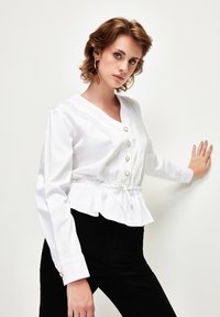 Camicia bianca con scollo a V, cordoncino nella parte inferiore e dettagli con bottoni dorati. Presenta decorazioni a pintuck verticali e maniche lunghe. Abbinata a pantaloni neri.