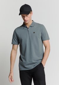 Grijze polo met korte mouwen, een zip-kraag en een klein zwart logo, gecombineerd met een zwarte pet en broek. Gladde textuur.